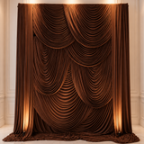 A large draped brown velvet curtain backdrop with layered scalloped folds, displayed in an elegant cream-colored hall with warm floor and wall lighting.