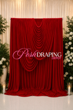 A deep red velvet photography backdrop curtain with elegant draped folds, shown as a product display. The backdrop is flanked by decorative green foliage and white flowers, with a 'Posh DRAPING DEPOT' watermark overlay in the center.